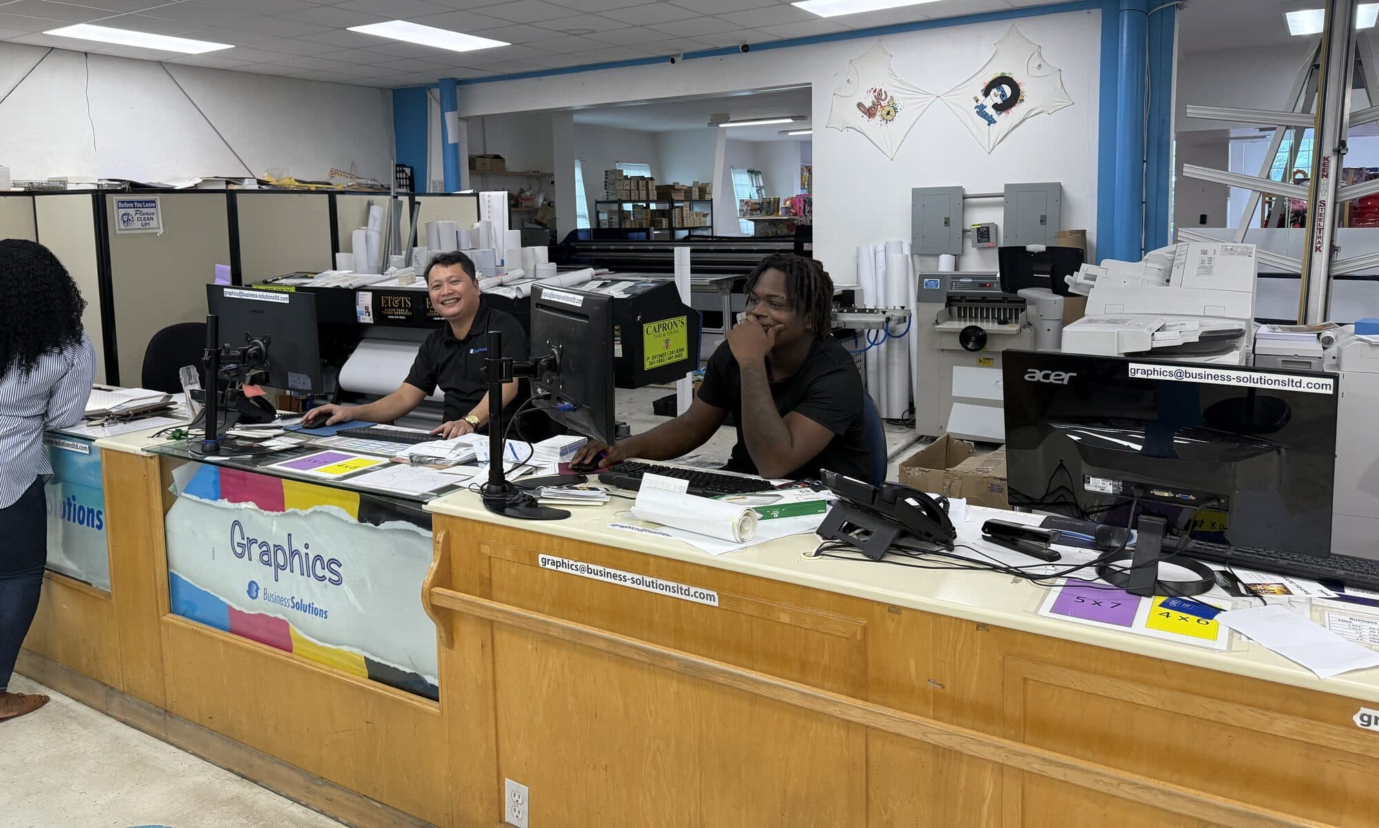 The graphics counter at Business Solutions in Providenciales: two designers working at the desk, with plan printers, rolls of media and a guillotine behind them.